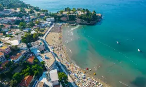 Widok na plażę w Czarnogórze z mnóstwem parasoli i kąpiącymi się ludźmi. W tle widać miasteczko i turkusowe morze. Idealne miejsce na wakacje z dziećmi.