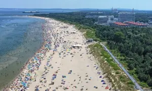 Tłum ludzi wypoczywa na najszerszej plaży w Polsce. W tle widać morze z statkiem i zalesione wybrzeże z nowoczesnymi budynkami.