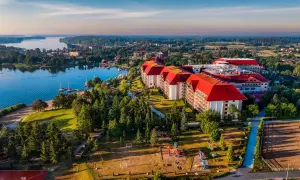 Hotel Gołębiewski w Mikołajkach, z czerwonymi dachami, otoczony zielenią i jeziorem. Rok budowy hotelu to 2004.