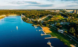 Malownicze jezioro z piaszczystą plażą, pomostami i terenami zielonymi, otoczone lasem i zabudowaniami miejskimi.