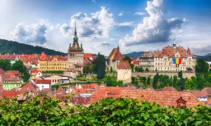 Widok na zabytkowe miasto z wieżą zegarową i flagą Rumunii, na tle zielonych wzgórz i błękitnego nieba.