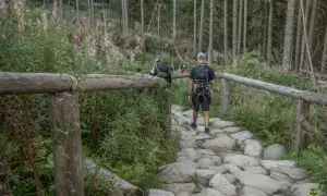 Kamienista ścieżka w lesie z drewnianym ogrodzeniem i turystami idącymi w kierunku gór.