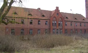 Opuszczony budynek z cegły, zniszczone okna i dach, otoczony dziką roślinnością. Idealne miejsce dla poszukiwaczy nawiedzonych miejsc w Polsce.