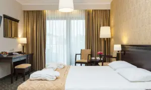 Elegancki pokój hotelowy z łóżkiem, biurkiem i fotelami. W tle zasłony i okno. Hotel Lambert historia w każdym detalu.