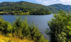 Malownicze góry i spokojne jezioro w słoneczny dzień. W oddali widać domy na zboczu, a na pierwszym planie bujną zieleń.
