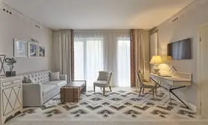 Elegancki salon w hotelu 5 gwiazdkowym. Sofa, stolik, fotele i biurko tworzą przytulną przestrzeń. Wzory na dywanie i zasłonach dodają charakteru.