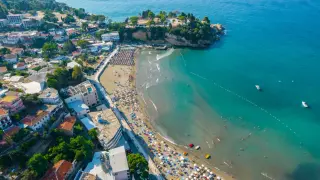 Widok na plażę w Czarnogórze z mnóstwem parasoli i kąpiącymi się ludźmi. W tle widać miasteczko i turkusowe morze. Idealne miejsce na wakacje z dziećmi.