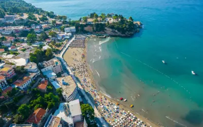 Widok na plażę w Czarnogórze z mnóstwem parasoli i kąpiącymi się ludźmi. W tle widać miasteczko i turkusowe morze. Idealne miejsce na wakacje z dziećmi.