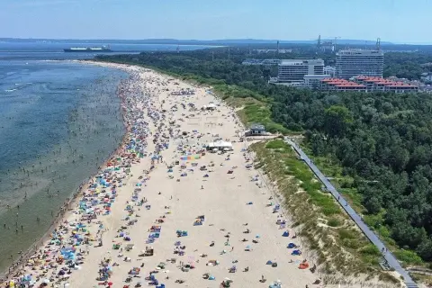 Tłum ludzi wypoczywa na najszerszej plaży w Polsce. W tle widać morze z statkiem i zalesione wybrzeże z nowoczesnymi budynkami.