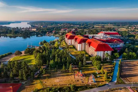 Hotel Gołębiewski w Mikołajkach, z czerwonymi dachami, otoczony zielenią i jeziorem. Rok budowy hotelu to 2004.