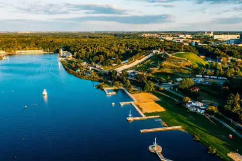 Malownicze jezioro z piaszczystą plażą, pomostami i terenami zielonymi, otoczone lasem i zabudowaniami miejskimi.