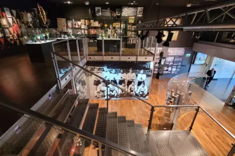 Wnętrze muzeum sportu w Warszawie. W gablotach pamiątki, puchary i zdjęcia. Schody prowadzą do kolejnych ekspozycji.