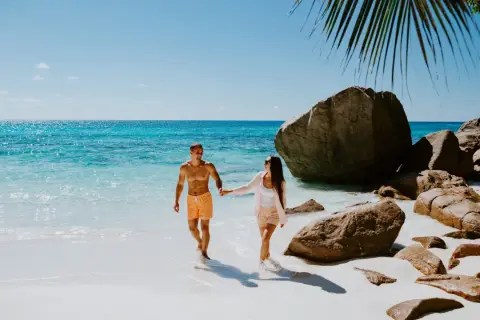 Para idealny czas na Seszele, kiedy jechać? Para spaceruje po rajskiej plaży z turkusową wodą i granitowymi skałami.