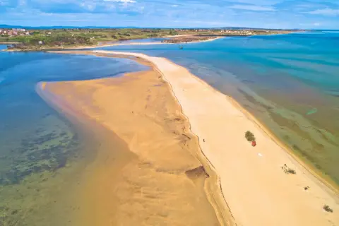 Piaszczyste plaże Chorwacji w całej okazałości. Długi, piaszczysty cypel otoczony turkusową wodą, z widokiem na miasteczko w oddali.