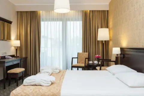 Elegancki pokój hotelowy z łóżkiem, biurkiem i fotelami. W tle zasłony i okno. Hotel Lambert historia w każdym detalu.