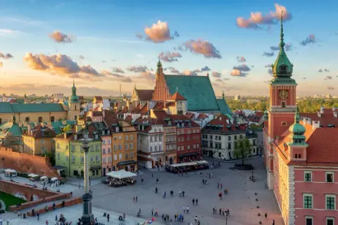 Zachód słońca nad Zamkiem Królewskim i Rynkiem Starego Miasta w Warszawie. Kolorowe kamienice, wieża zegarowa i chmury na niebie tworzą malowniczy widok.