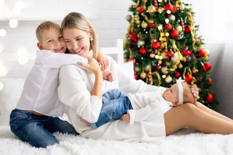 Rodzina cieszy się świętami Bożego Narodzenia w hotelu. Mama tuli synka, a córeczka śpi na jej kolanach. W tle choinka.