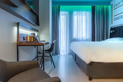 Nowoczesny pokój w Radisson Park Hotel Poznań: wygodne łóżko, biurko z czajnikiem i fotelem, zasłony i inspirujący cytat na ścianie.