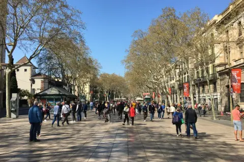 Tłum ludzi spaceruje po słynnej alei La Rambla w Barcelonie, otoczonej drzewami i budynkami.