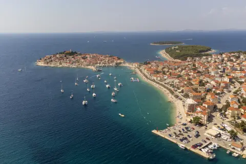 Malownicze miasteczko na wybrzeżu Chorwacji, gdzie turkusowe morze otacza wyspę z czerwonymi dachami. Wiele łodzi cumuje przy brzegu, a plaża tętni życiem.