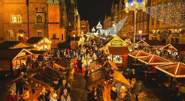 Tłoczny wrocławski jarmark bożonarodzeniowy nocą, rozświetlony tysiącami lampek. Ludzie spacerują między drewnianymi straganami, ciesząc się świąteczną atmosferą.