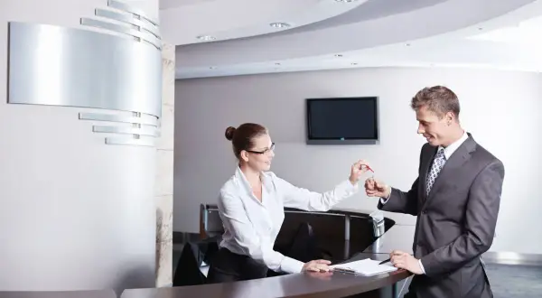 Miła technik hotelarstwa przekazuje klucze gościowi przy recepcji.