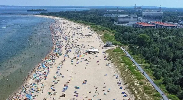 Tłum ludzi wypoczywa na najszerszej plaży w Polsce. W tle widać morze z statkiem i zalesione wybrzeże z nowoczesnymi budynkami.