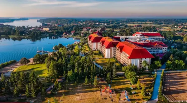 Hotel Gołębiewski w Mikołajkach, z czerwonymi dachami, otoczony zielenią i jeziorem. Rok budowy hotelu to 2004.