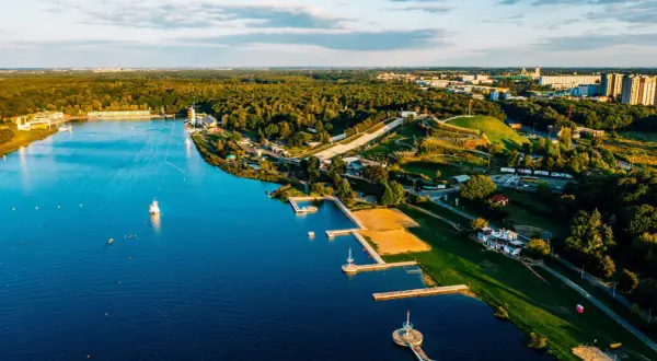Malownicze jezioro z piaszczystą plażą, pomostami i terenami zielonymi, otoczone lasem i zabudowaniami miejskimi.