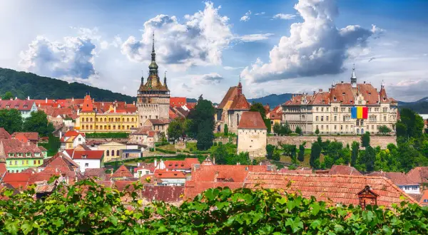 Widok na zabytkowe miasto z wieżą zegarową i flagą Rumunii, na tle zielonych wzgórz i błękitnego nieba.