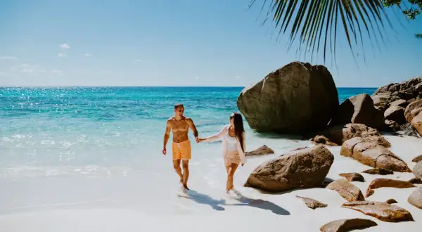Para idealny czas na Seszele, kiedy jechać? Para spaceruje po rajskiej plaży z turkusową wodą i granitowymi skałami.