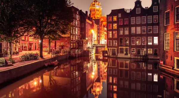 Amsterdam nocą, kanał z odbiciem zabytkowych kamienic i kościoła. Czerwona ulica Amsterdamu rozświetlona latarniami.