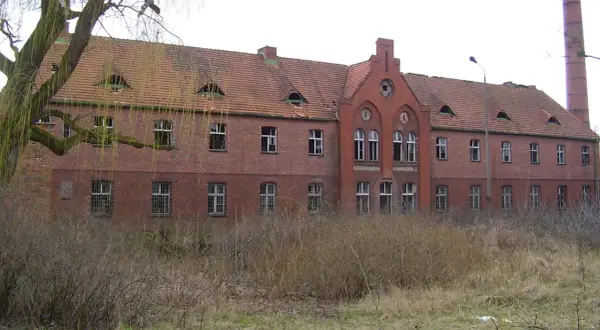 Opuszczony budynek z cegły, zniszczone okna i dach, otoczony dziką roślinnością. Idealne miejsce dla poszukiwaczy nawiedzonych miejsc w Polsce.