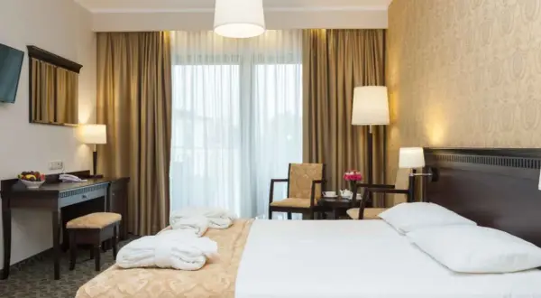 Elegancki pokój hotelowy z łóżkiem, biurkiem i fotelami. W tle zasłony i okno. Hotel Lambert historia w każdym detalu.