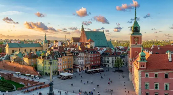 Zachód słońca nad Zamkiem Królewskim i Rynkiem Starego Miasta w Warszawie. Kolorowe kamienice, wieża zegarowa i chmury na niebie tworzą malowniczy widok.