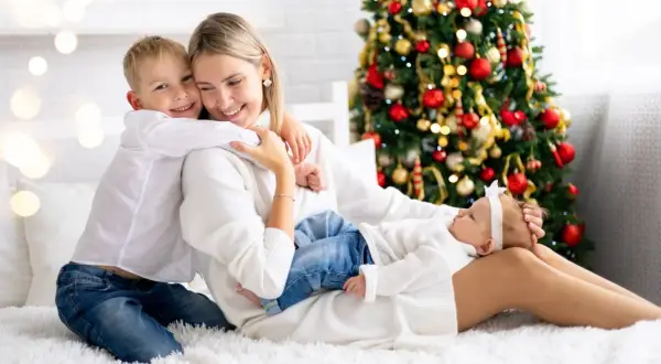 Rodzina cieszy się świętami Bożego Narodzenia w hotelu. Mama tuli synka, a córeczka śpi na jej kolanach. W tle choinka.