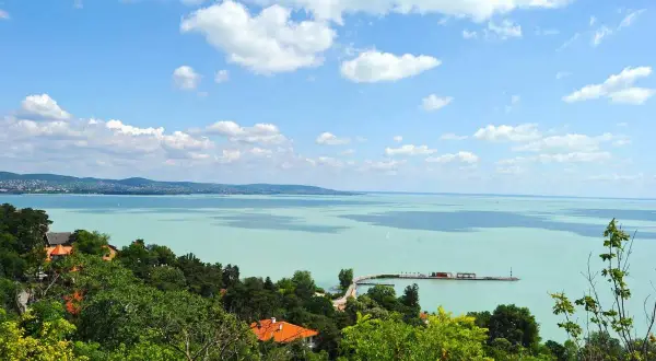 Widok na turkusowe wody Balatonu z zielonymi wzgórzami i domami. To jedno z najpiękniejszych miejsc nad Balatonem, z molo i żaglówkami na horyzoncie.