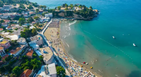 Widok z lotu ptaka na tętniące życiem czarnogórskie miasta turystyczne z plażą pełną parasoli i turystów, oraz turkusowym morzem.