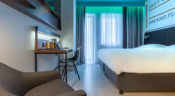 Nowoczesny pokój w Radisson Park Hotel Poznań: wygodne łóżko, biurko z czajnikiem i fotelem, zasłony i inspirujący cytat na ścianie.