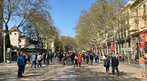 Tłum ludzi spaceruje po słynnej alei La Rambla w Barcelonie, otoczonej drzewami i budynkami.