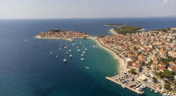 Malownicze miasteczko na wybrzeżu Chorwacji, gdzie turkusowe morze otacza wyspę z czerwonymi dachami. Wiele łodzi cumuje przy brzegu, a plaża tętni życiem.