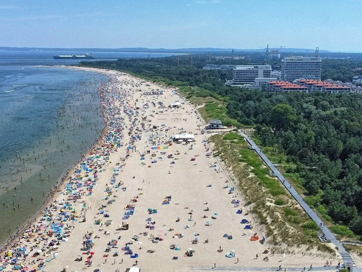 Tłum ludzi wypoczywa na najszerszej plaży w Polsce. W tle widać morze z statkiem i zalesione wybrzeże z nowoczesnymi budynkami.