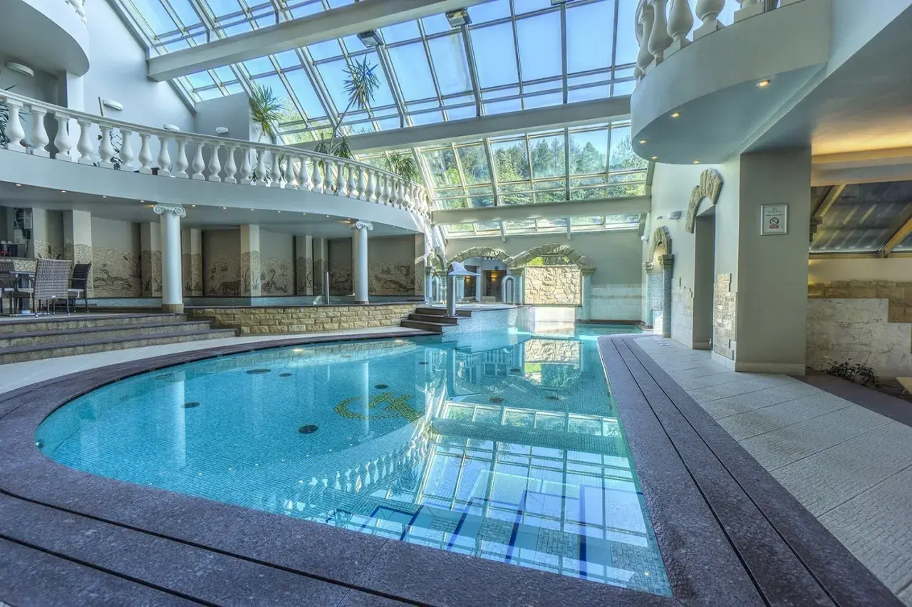 Basen w hotelu Szczyrk Klimczok z przeszklonym dachem, elegancką balustradą i mozaikami na ścianach.