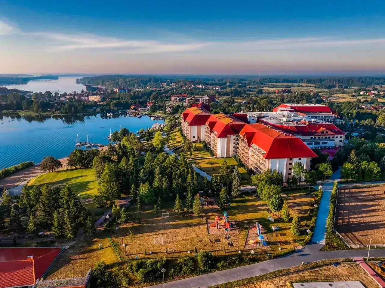 Hotel Gołębiewski w Mikołajkach, z czerwonymi dachami, otoczony zielenią i jeziorem. Rok budowy hotelu to 2004.