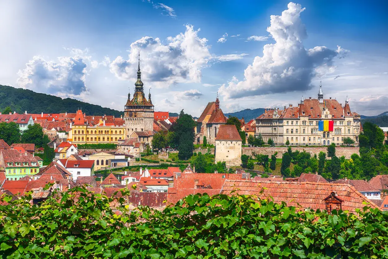 Widok na zabytkowe miasto z wieżą zegarową i flagą Rumunii, na tle zielonych wzgórz i błękitnego nieba.