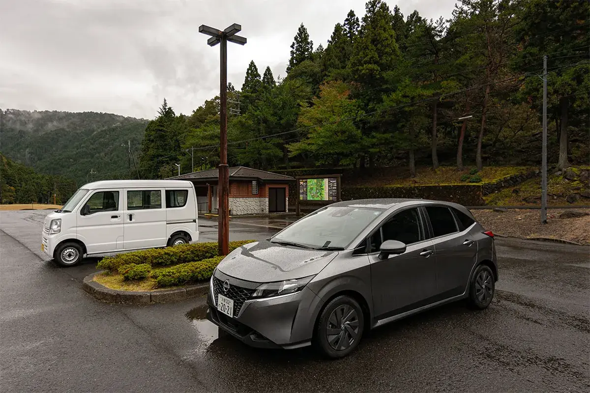Szary Nissan i biały van na parkingu w Japonii. Widać, jaki ruch jest w Japonii, gdy podróżuje się po malowniczych terenach.