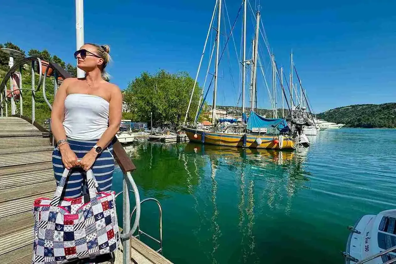 Kobieta z torbą w marynistycznym stylu na pomoście, w tle żaglówki i turkusowa woda. Idealny dzień na Chorwacji w maju.
