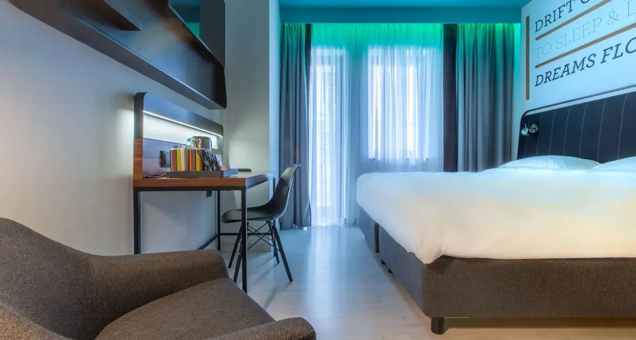 Nowoczesny pokój w Radisson Park Hotel Poznań: wygodne łóżko, biurko z czajnikiem i fotelem, zasłony i inspirujący cytat na ścianie.