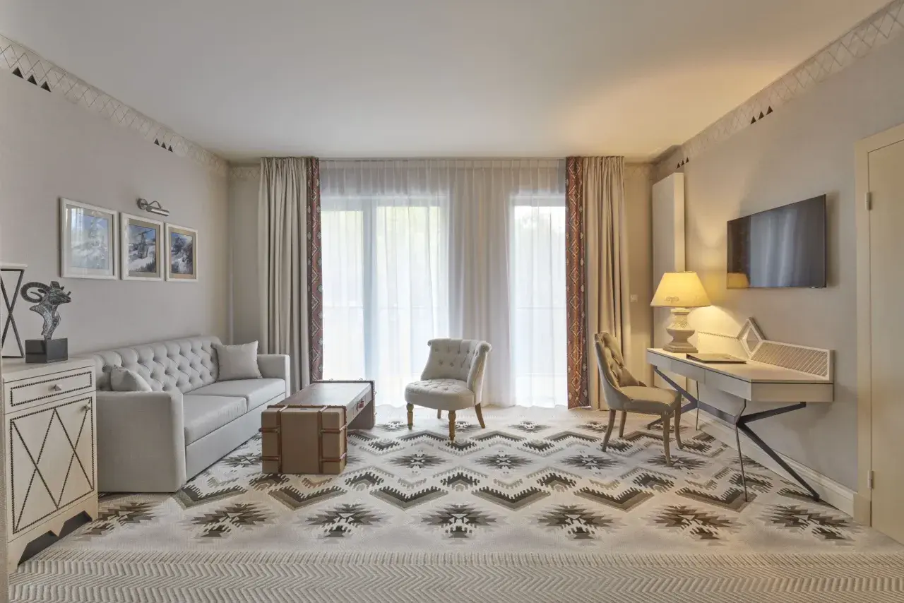 Elegancki salon w hotelu 5 gwiazdkowym. Sofa, stolik, fotele i biurko tworzą przytulną przestrzeń. Wzory na dywanie i zasłonach dodają charakteru.