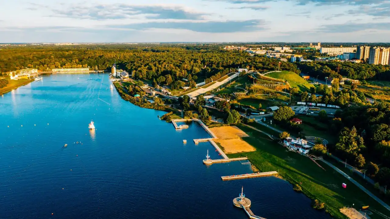 Malownicze jezioro z piaszczystą plażą, pomostami i terenami zielonymi, otoczone lasem i zabudowaniami miejskimi.